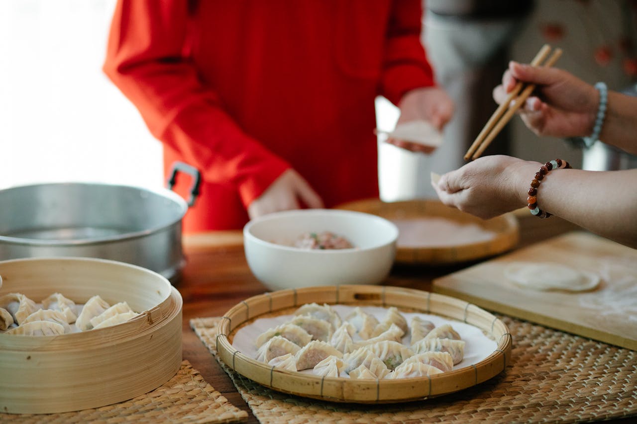 Two people preparing homemade dumplings on a bamboo tray in a warm kitchen setting.