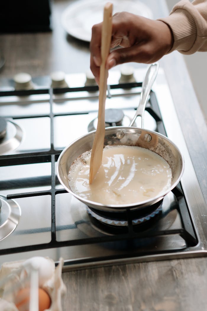 Person stirring sauce in frying pan on a gas stove, showcasing home cooking.